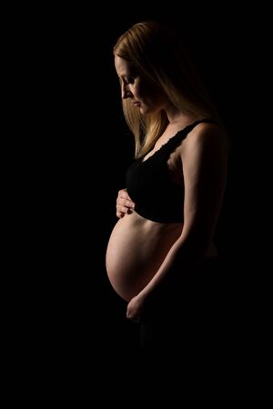 Pregnant woman in black underwear  holding belly on black background. Portrait of excited, happy, blond, young woman in pregnant studio shoot. Concept of maternity and expectation. Isolated black background.の写真素材