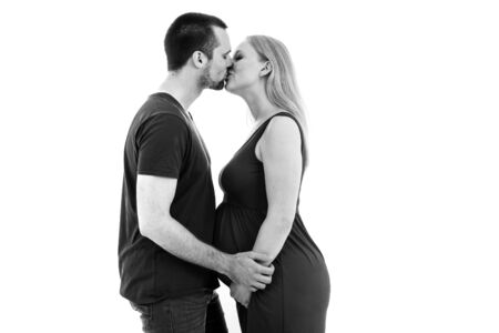 Young pregnant couple in love expecting child, studio portrait. Husband hugging his pregnant wife in intimate relationship. Young pregnant couple kissing and holding hands. Isolated white background.の写真素材
