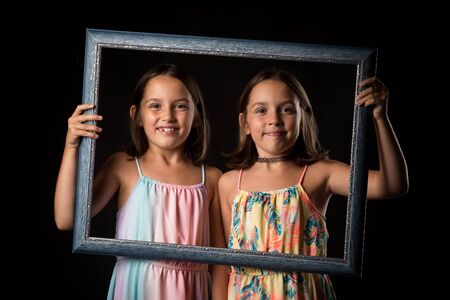 Identical twin girls are making happy expressions with picture frame. Children, sisters, girls posing in studio with picture frame, making different facial expressions. Family portrait, frontal view.の写真素材