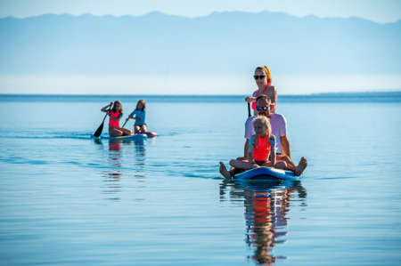 Happy Family on SUP stand up paddle on vacation. Active family riding SUP boards and paddling in the ocean on a beautiful morning. Mother and three girls share one SUP board with yachts in backgroundの写真素材