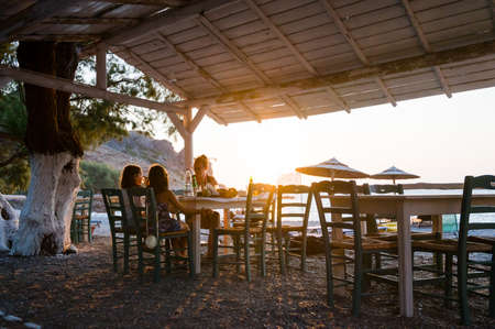 Family enjoying dinner in a restaurant on the beach at sunset. People in a cafeÂ on a beautiful beach tavern with sunset view of the Sfinari beach in Crete, Greece.の写真素材