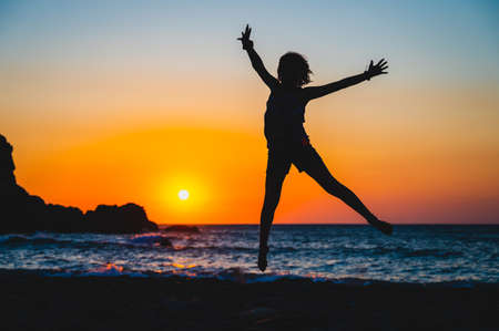 Silhouette of a child jumping in sunset on a beach. Little girl jumping with hands raisedÂ on a sunset beach with the sea in the background. Amazing sunset in Sfinari beach, Crete, Greece.の写真素材