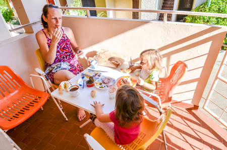 Family eating breakfast on vacation. Mother and children baby twinsÂ are eating breakfast - breadÂ  yogurt, ham, cheese on a balcony or terrace on a sunny morning on holidays.の写真素材