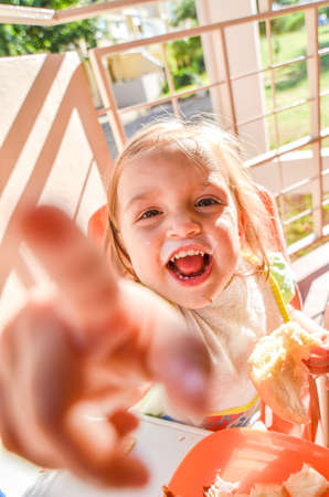 Happy baby eating breakfast on vacation. Child with a dirty mouth and bib is eating bread and yogurt on a balcony or terrace on a sunny morning on holidays.の写真素材
