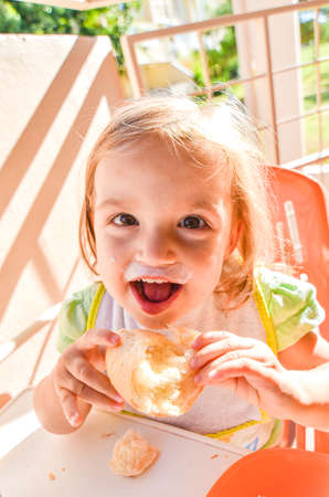 Happy baby eating breakfast on vacation. Child with a dirty mouth and bib is eating bread and yogurt on a balcony or terrace on a sunny morning on holidays.の写真素材