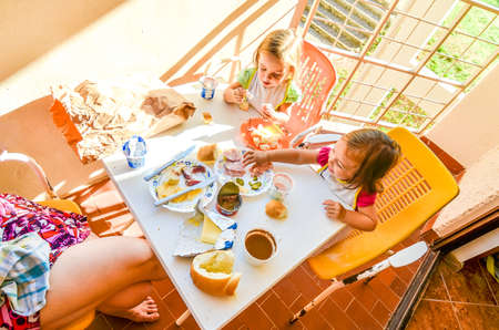 Family eating breakfast on vacation. Mother and children baby twinsÂ are eating breakfast - breadÂ  yogurt, ham, cheese on a balcony or terrace on a sunny morning on holidays.の写真素材