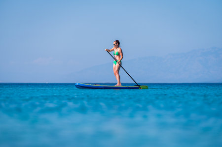 Woman riding SUP stand up paddle on vacation. Active female riding SUP boards and paddling in the ocean on sunny day. Athletic woman on SUP board with yachts in background, Silba, Croatia.の写真素材