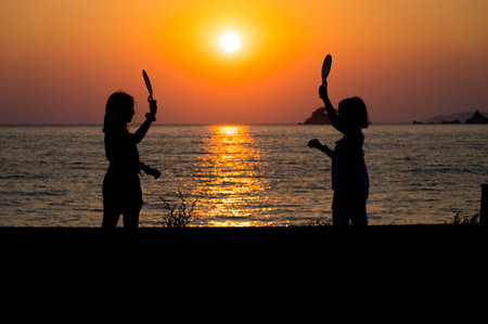Silhouette of children playing tennis with sunset sun on a beach. Little girls playing on a sunset beach with the sea in the background. Amazing sunset in the Adriatic sea.の写真素材