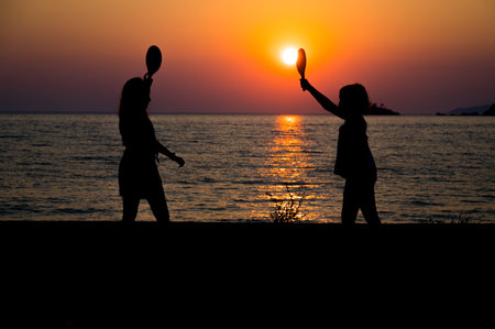 Silhouette of children playing tennis with sunset sun on a beach. Little girls playing on a sunset beach with the sea in the background. Amazing sunset in the Adriatic sea.の写真素材