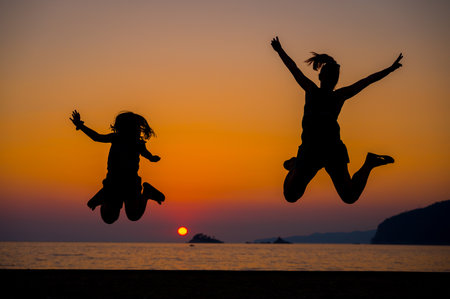 Silhouette of mother and children jumping at sunset on a beach. People jumping with hands raised on a sunset beach with the sea in the background. Amazing sunset in the Adriatic sea.の写真素材