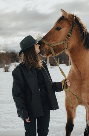 cowgirl kissing her horse in the snow with hat and leather jacket on a cloudy dayの写真素材
