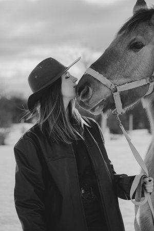 cowgirl kissing her horse in the snow with hat and leather jacket on a cloudy day