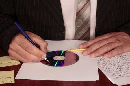 a man in suit and tie is writing on the compact disk, we can see only his hands, disk, pen and some...