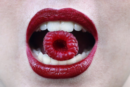 A woman with red lips holds a raspberry between her teethの写真素材