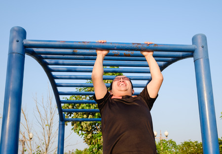 fitness ,sport, exercise, training and lifestyle concept - one young adult fat man doing pull ups on horizontal bar outdoors for   losing weight in eveningの写真素材