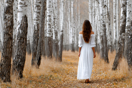 Woman in white dress walking through a birch forest with autumn leavesの素材