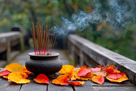 Incense sticks burning in a censer, surrounded by colorful autumn leaves on a wooden surfaceの素材