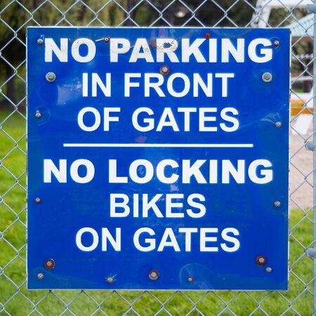 A sign attached to a chain-link fence that reads: NO PARKING IN FRONT OF GATES / NO LOCKING BIKES ON...