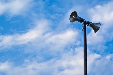 A pair of loudspeaker megaphones mounted atop a tall pole set against a cloudy blue sky.の写真素材