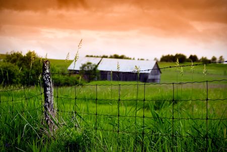 An old, rotting wire fence stands in the foreground with an old barn in the background, under a stormy, orange sky.の写真素材