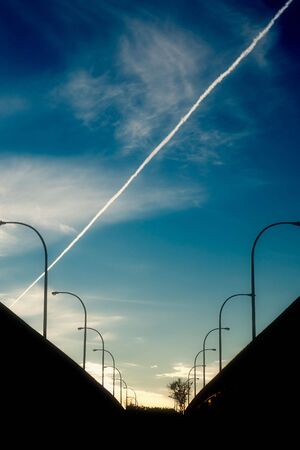 An airliner contrail cuts through a dynamic, cloudy sky with the silhouettes of streetlights lining a highway bridge in the foreground.の写真素材