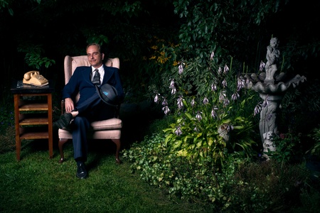 A man wearing a suit sits on a chair beside a an old style rotary telephone in a lush, green garden の写真素材