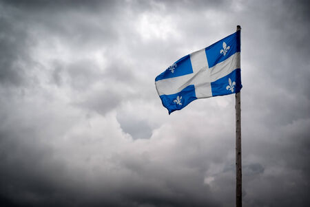 The flag of Quebec   FleurdelisÃ©   waves in the wind in front of the tumultuous clouds of stormy skies の写真素材