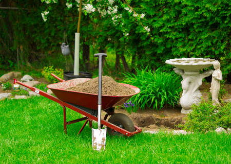 A red Wheelbarrow sits full of dirt in a lush green garden as spring gardening work is underway.の写真素材