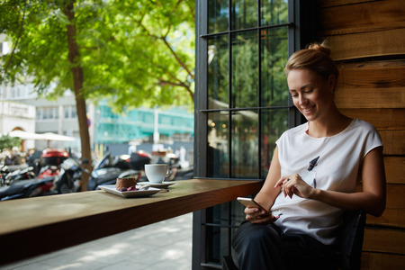 Charming woman with beautiful smile reading good news on mobile phone during rest in coffee shop, happy Caucasian female watching her photo on cell telephone while relaxing in cafe during free time Charming woman with beautiful smile reading good news on mobile phone during rest in coffee shop, happy Caucasian female watching her photo on cell telephone while relaxing in cafe during free time