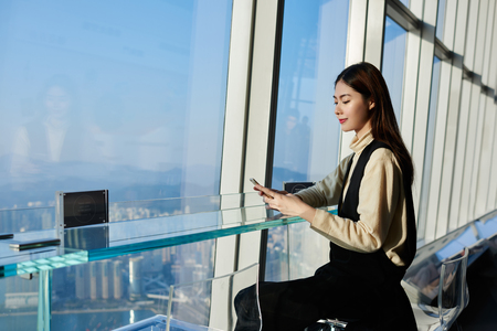 Woman successful lawyer is reading news in internet via cell telephone, while is waiting her client in modern interior with metropolitan city outside window. Young Japanese female is chatting on phone Woman successful lawyer is reading news in internet via cell telephone, while is waiting her client in modern interior with metropolitan city outside window. Young Japanese female is chatting on phone