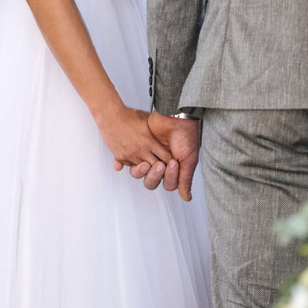 Back view of a bride in white dress and groom in a suit holding each other's hands in the church during a ceremony, square imageの写真素材