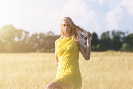 Blonde girl posing in the green barley field and enjoying summer in nature golden hour sunset. Girl in a yellow dress.の写真素材