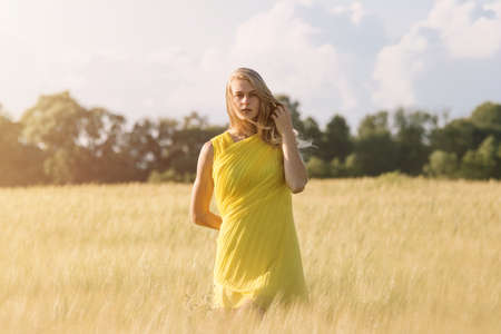 Blonde girl posing in the green barley field and enjoying summer in nature golden hour sunset. Girl in a yellow dress.の写真素材