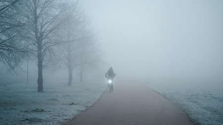 View of men cycling on road in the morning fog. A man rides a bike with a turned on headlight in deep fog on the promenade near the frozen treesの写真素材