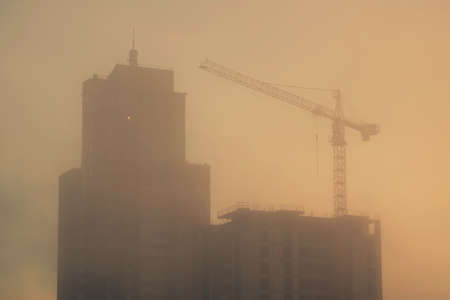 Silhouette of a construction crane and unfinished residential building on a foggy autumn morning during the sunrise. Housing construction, apartment blockの写真素材