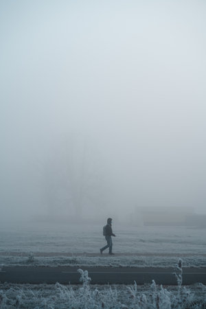 Young man with face mask walking alone on sidewalk in deep fog during the cold autumn morning with the frost. Side viewの写真素材