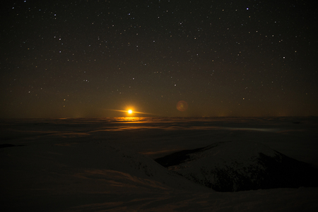 The moon rises from the horizon behind the mountains covered with clouds. Cosmic landscape, over the cloudsの写真素材