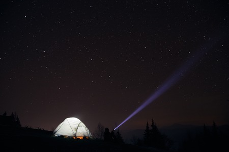 tourist with flashlight near his camp tent under a sky full of stars, night mountain lonelinessの写真素材