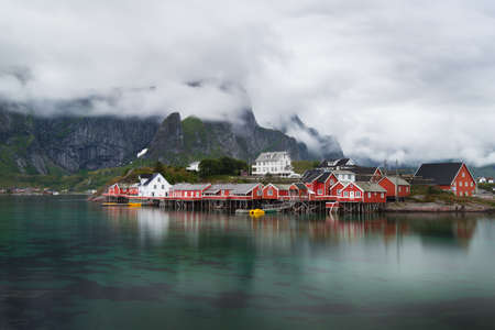 Amazing view of Reine - fishing village with red houses near green fjord and mountains in clouds after rain. Beauty of Hamnoya, Lofoten islands, Norwayの写真素材