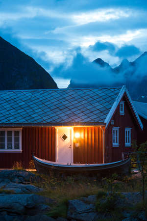 typical Norway red fishing hut with fishing wooden boat in the evening near great mountainsの写真素材