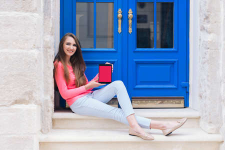 Beautiful young blonde female student sitting with tablet  on the stairs in front of blue doors.の写真素材