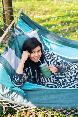 Happy healthy eating girl drinking green smoothie detox outdoors in hammock. Woman on weight loss diet vegan nutrition cleanse.の写真素材
