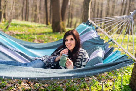 Happy healthy eating girl drinking green smoothie detox outdoors in hammock. Woman on weight loss...
