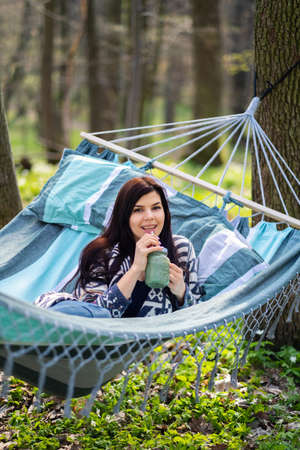 Happy healthy eating girl drinking green smoothie detox outdoors in hammock. Woman on weight loss diet vegan nutrition cleanse.の写真素材