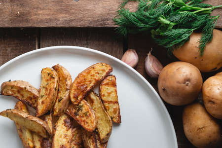 Delicious baked country potatoes in a large white plate with spices, dill and green onions, on a wooden tableの写真素材
