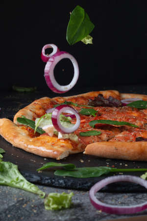 Traditional Italian pepperoni pizza with tomatoes, cheese and flying onions and herbs, on a table of dark slate on a black backgroundの写真素材