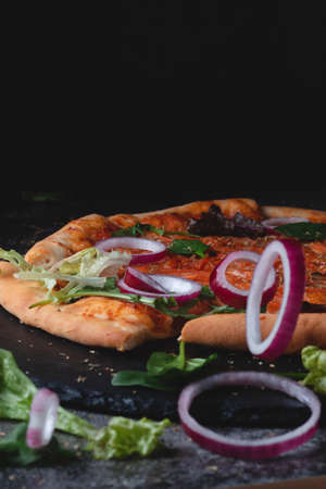 Traditional Italian pepperoni pizza with onions, herbs, tomatoes and cheese on a dark slate table and black backgroundの写真素材