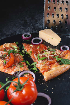 Slices Traditional Italian pepperoni pizza with tomatoes, grated metal cheese, onions and herbs, on a table of dark slate on a black backgroundの写真素材
