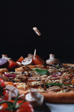 Pieces of flying fall mushrooms on Homemade Italian pizza on a cutting wooden board with ham, tomatoes, cheese, onions and herbs lies on a dark stone slate tableの写真素材