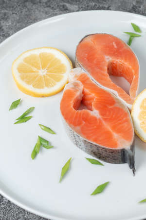 Juicy fresh salmon steak in a white plate on a wooden cutting board with onion, lemon and spices on a natural slate stone table, close-up. Deliciously cooked salmon fillet.の写真素材